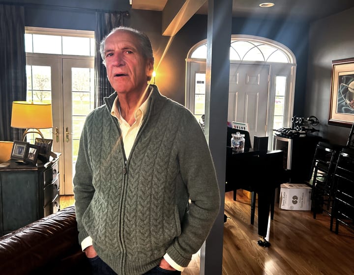 In a warmly lit living room, an older man stands beside a piano, embodying a moment of reflection and tranquility.