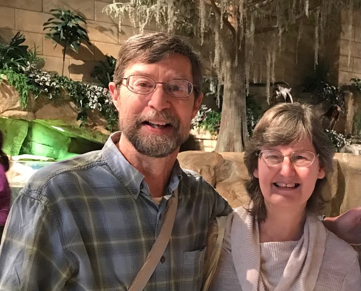 A smiling man and woman pose together indoors in front of a nature-themed backdrop with rocks, plants, and trees.
