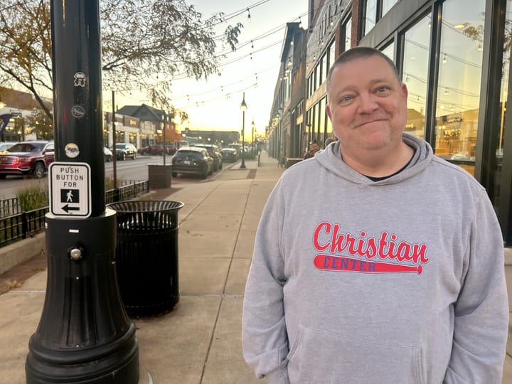 A middle-aged man in a hoodie sweatshirt stands on a city street in late afternoon.