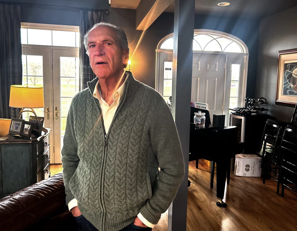 In a warmly lit living room, an older man stands beside a piano, embodying a moment of reflection and tranquility.