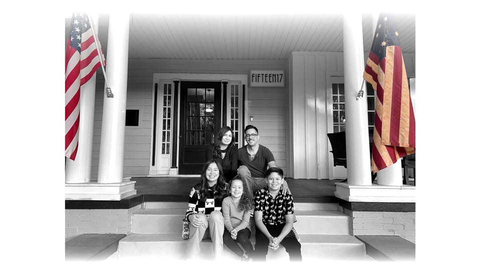 A smiling family of a woman and a man with their three children sit on the steps of an older well-kept home flanked by American flags.
