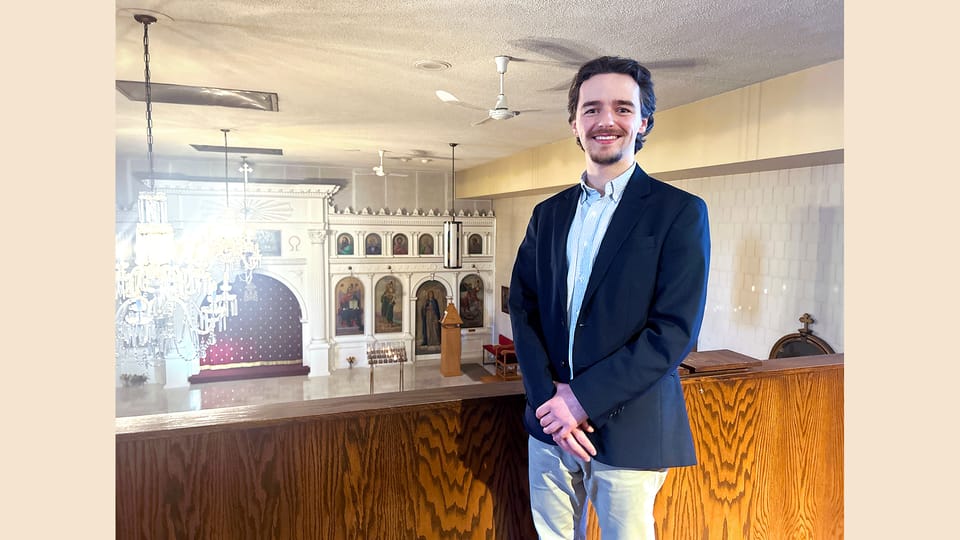 A young man in a blue blazer stands smiling in the balcony of a Greek Orthodox church.